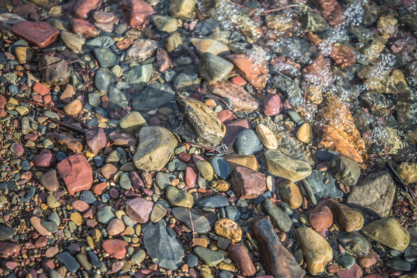 Fine Glacier National Park photography print of the calm shores gently rolling over multi-colored stones along the lakeshore.