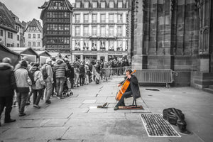 Fine black and white photographic and art print of a solo colorful cellist playing outside the doors of Strasbourg Cathedral as Christmas crowds gather and listen as they wait to enter.